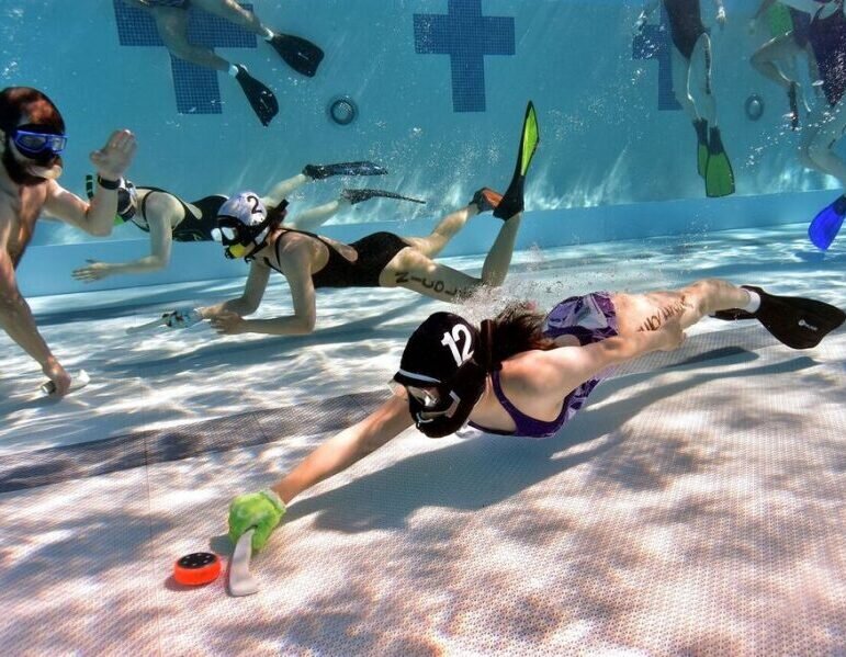 An underwater hockey player in a green glove stretching toward an orange puck on the pool floor
